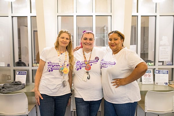 Three clinic staffers pose near front desk in t-shirts reading “Comprehensive Healthcare since 1976.”