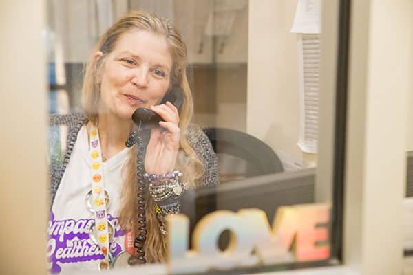 A staff member wearing a “Compassionate Healthcare since 1976” t-shirt smiles while holding a phone to their ear at the front desk of the clinic.