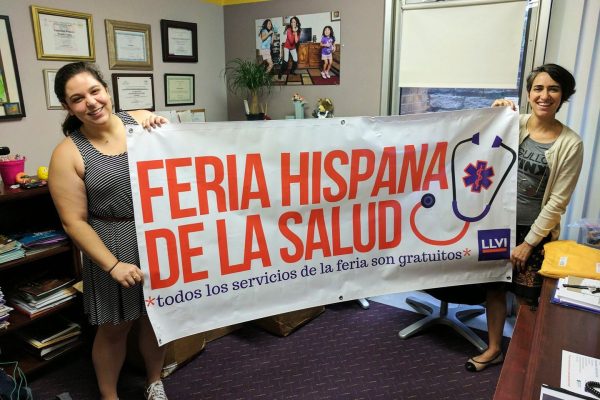 Two volunteers standing in an office holding up a banner that says "Feria Hispana de la Salud"