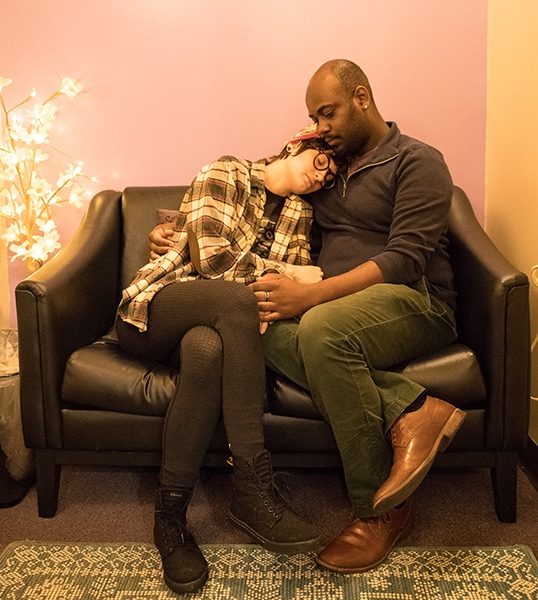 Models sit on a black leather couch inside the clinic, the model on the right poses as if they are comforting the model on the left.