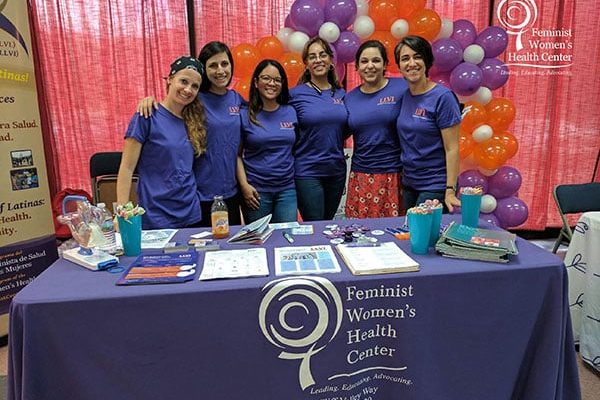 Members of the Lifting Latinx Voices Initiative team pose behind a Feminist Women’s Health Center table at an event.