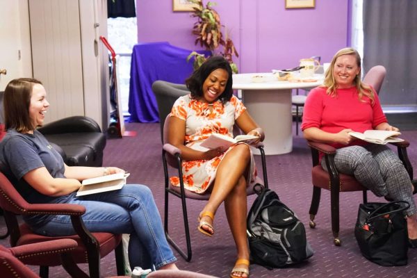 3 volunteers sit in chairs in a row holding books and smiling.